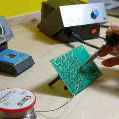 Precyzyjne lutowanie aluminium na płytce drukowanej. Dłoń trzymająca lutownicę, obok stojak i rolka cyny.