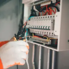 Elektryk w białych rękawiczkach pracuje przy skrzynce z bezpiecznikami. To kluczowy element zabezpieczenia przedlicznikowego.