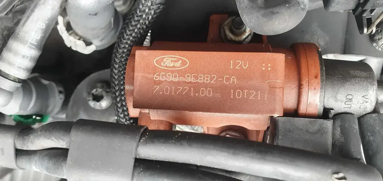 Zawór podciśnienia turbiny Ford Focus 1.6 TDCI, o numerze 6G90-9E882-CA, znajduje się w pobliżu przewodów i innych elementów silnika.